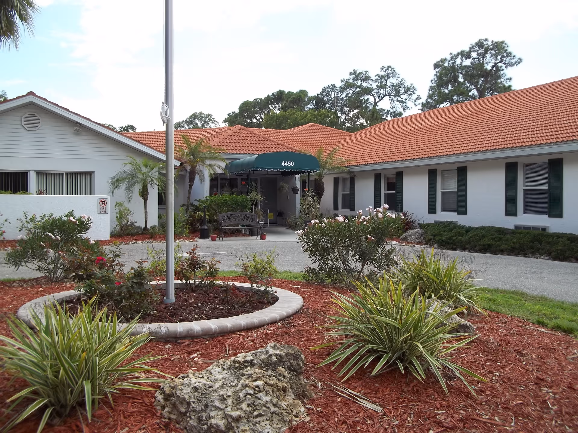 Exterior view of a single-story building with a red tiled roof and white walls, surrounded by landscaped garden beds with various plants and a flagpole in the center. The entrance has a green awning with the number 4450 displayed above it.