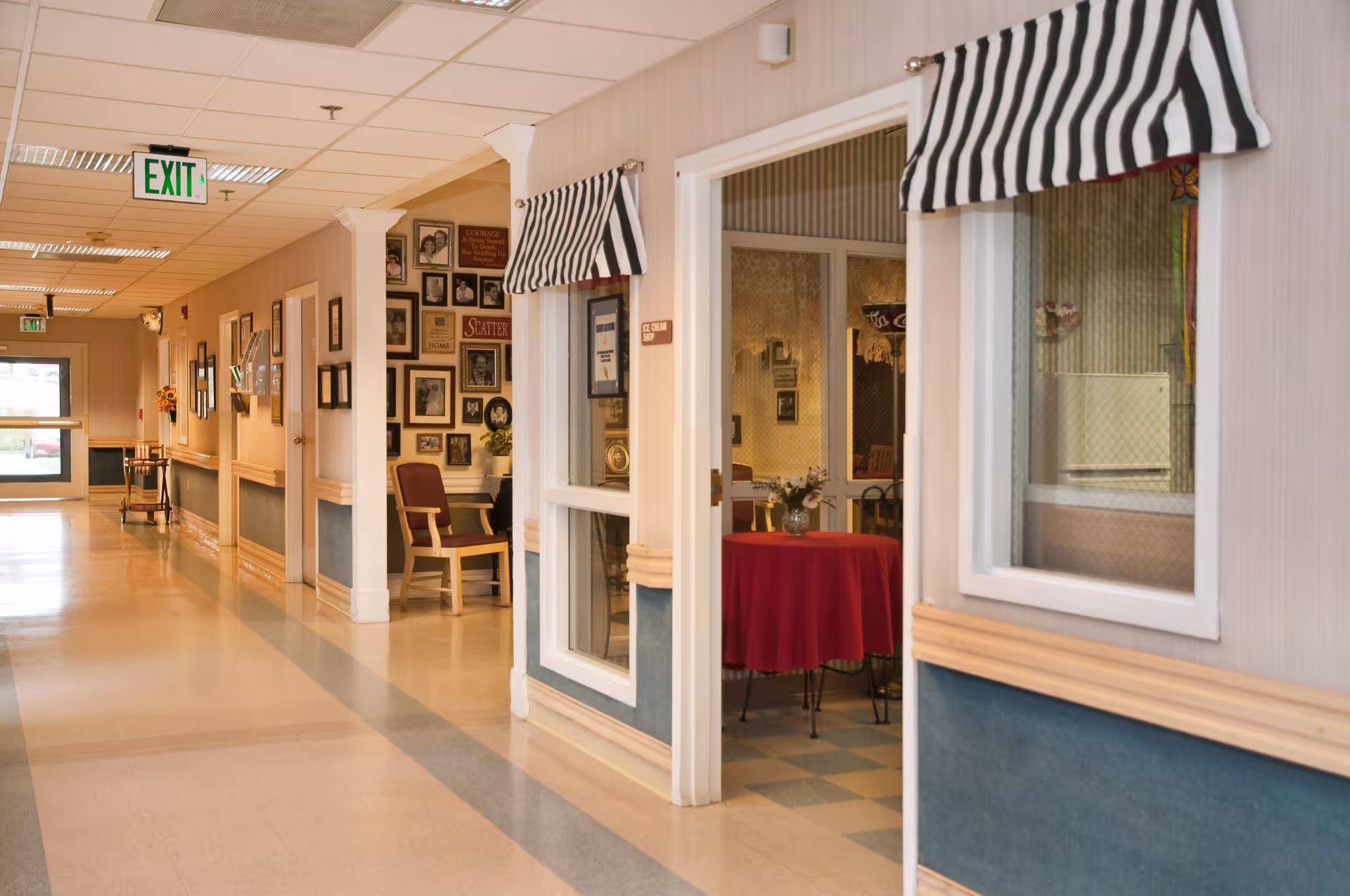 A bright interior hallway of a senior living facility with framed photos on the wall, seating, and a side room with a red-covered table visible through windows.