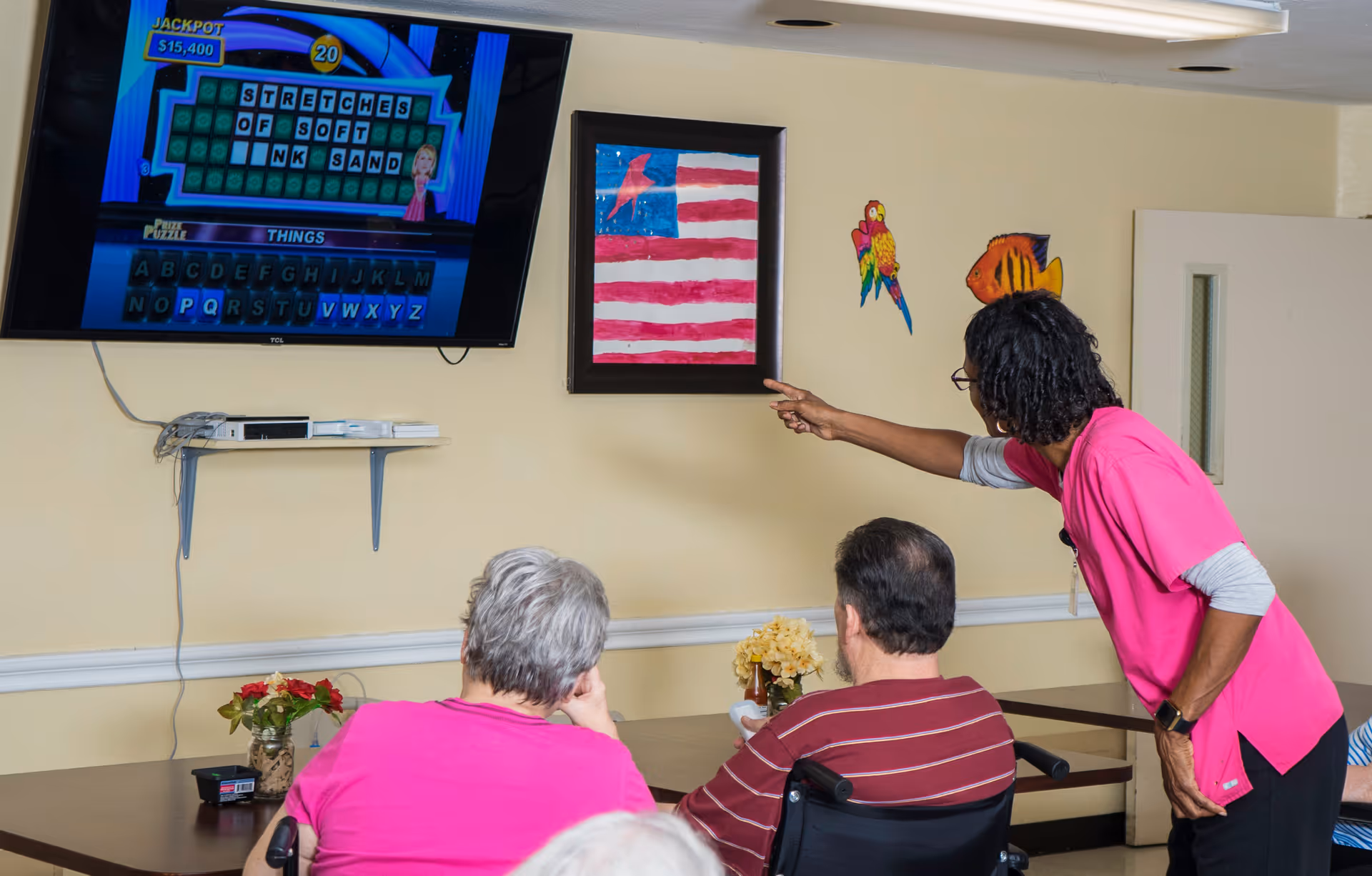 A caregiver in a pink uniform points at a wall-mounted TV screen displaying a word puzzle game while two elderly individuals, one in a wheelchair, watch attentively in a common room with beige walls decorated with a framed American flag and colorful fish and bird wall decals.