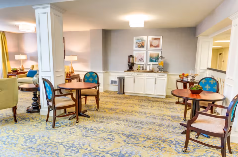 A bright and spacious common area with patterned carpet, round wooden tables, and upholstered chairs with blue and gold patterned backs. There are two white pillars, a sideboard with beverage dispensers, and framed artwork on the wall. The room is softly lit with ceiling lights and table lamps, and there is a seating area with a green sofa and a round coffee table in the background.