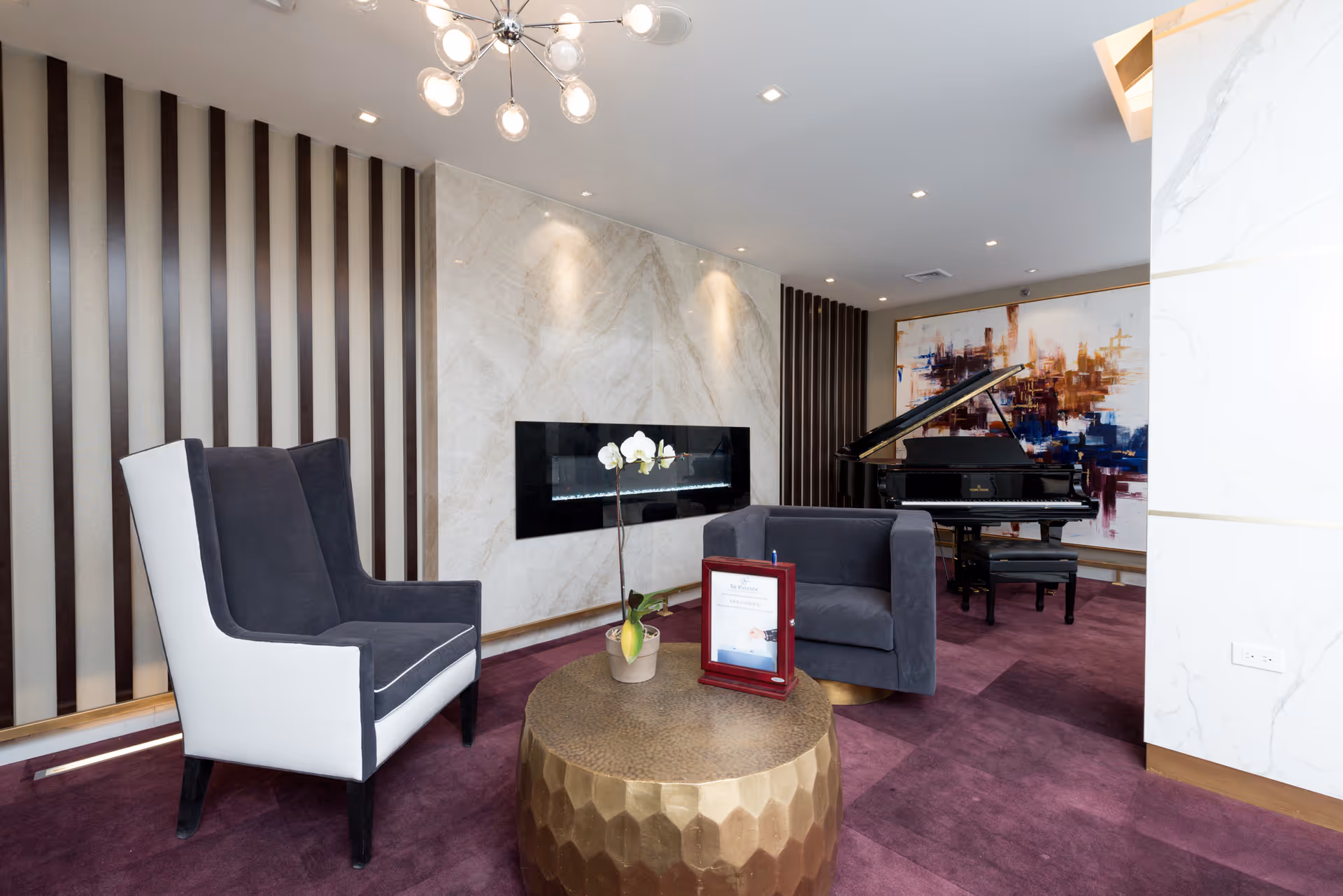 A modern lounge area with a dark gray armchair and a matching two-seater sofa arranged around a round gold coffee table with a potted orchid and a framed document on it. Behind the seating is a wall with vertical dark wood slats and a built-in rectangular electric fireplace set in a marble wall. In the background, there is a black grand piano in front of a large abstract painting with blue, orange, and white colors. The floor is covered with a purple carpet, and a contemporary chandelier with multiple exposed bulbs hangs from the ceiling.
