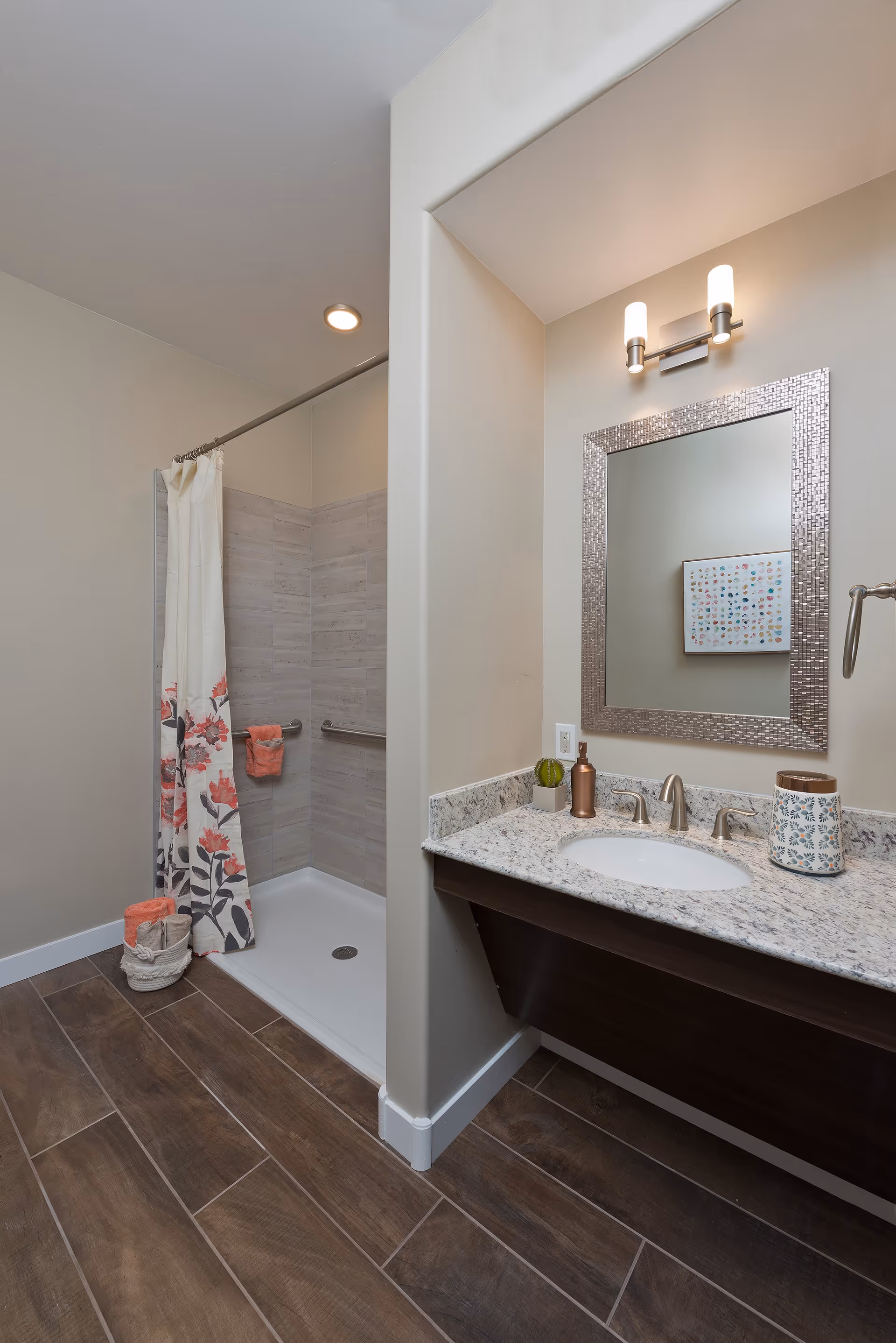A modern bathroom featuring a walk-in shower with a floral shower curtain and grab bars, a granite countertop with an under-mount sink, a decorative mirror with a textured frame, and a wall-mounted light fixture above the mirror. The floor is covered with wood-look tiles, and there are neatly folded towels in a basket near the shower.