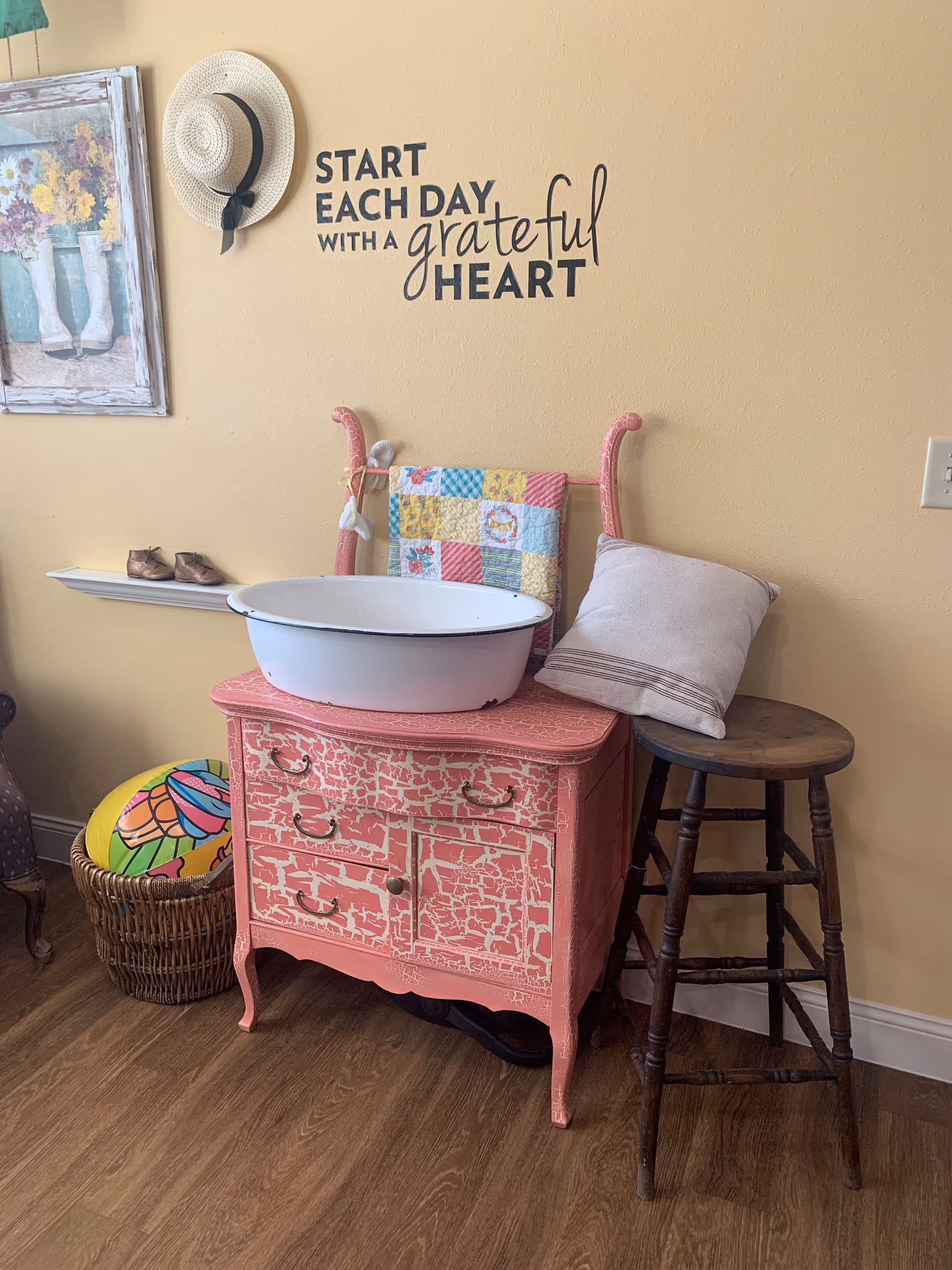 Decorative interior corner with a distressed pink dresser holding a white basin, a wooden stool, quilt, hat on the wall and a wall decal that says "Start each day with a grateful heart."