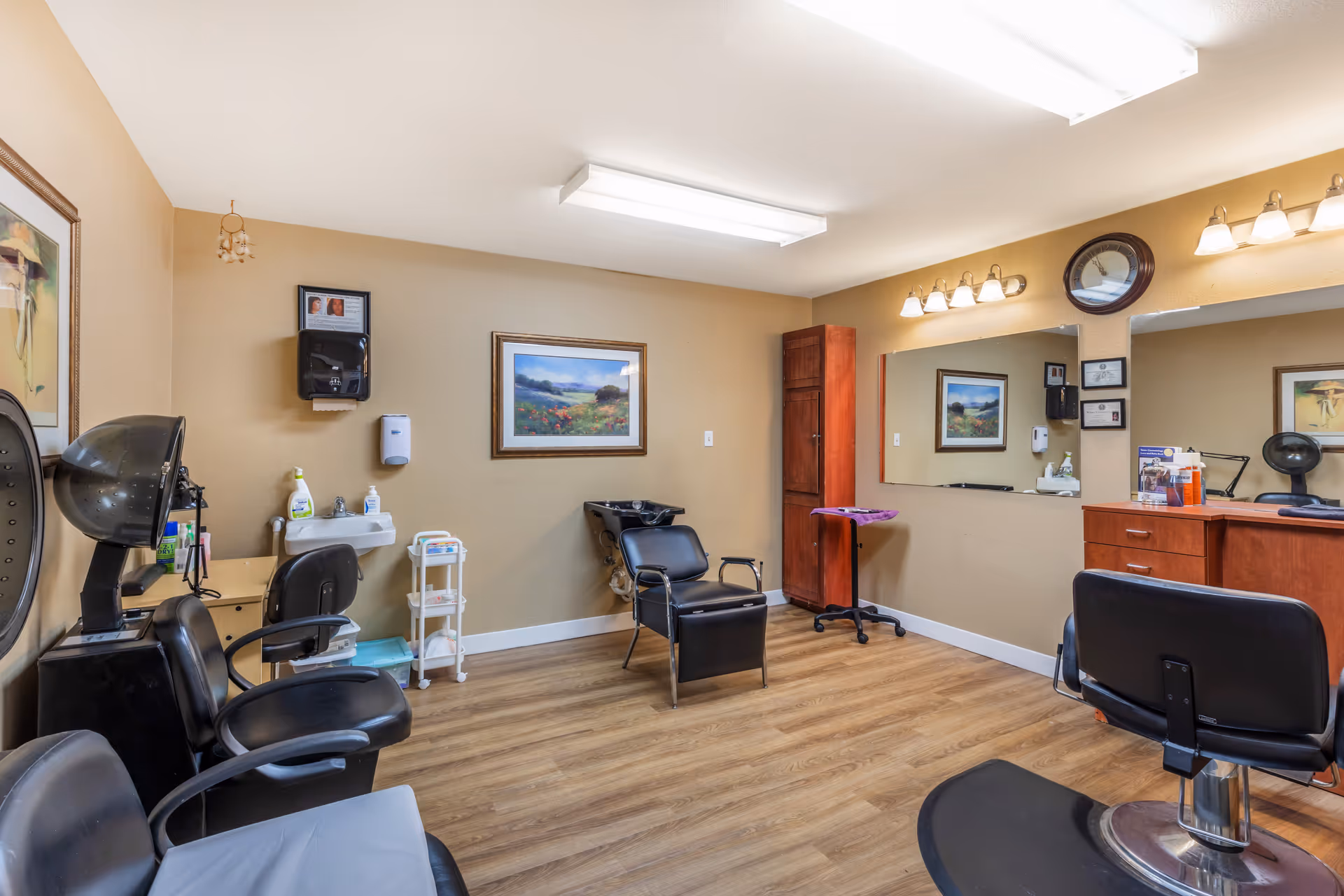 Interior view of a hair salon area in a senior living facility with multiple black salon chairs, a hair dryer, a sink for washing hair, a large mirror, wooden cabinets, and framed artwork on beige walls.