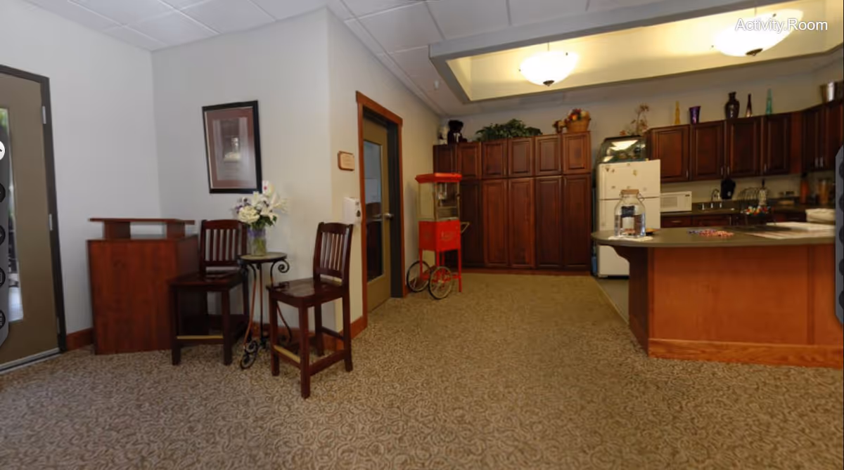 Interior view of an activity room with a carpeted floor, two wooden chairs and a small round table with a flower vase on it, a wooden podium, a popcorn machine, and a kitchen area with wooden cabinets, a refrigerator, microwave, and countertop. The ceiling has recessed lighting and there is a door with a glass panel.