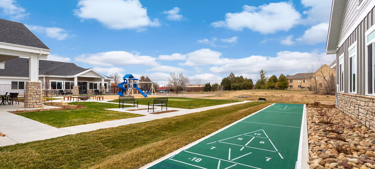 Outdoor courtyard at Greeley Village showing a shuffleboard court, benches, a playground, and surrounding buildings under a blue sky.
