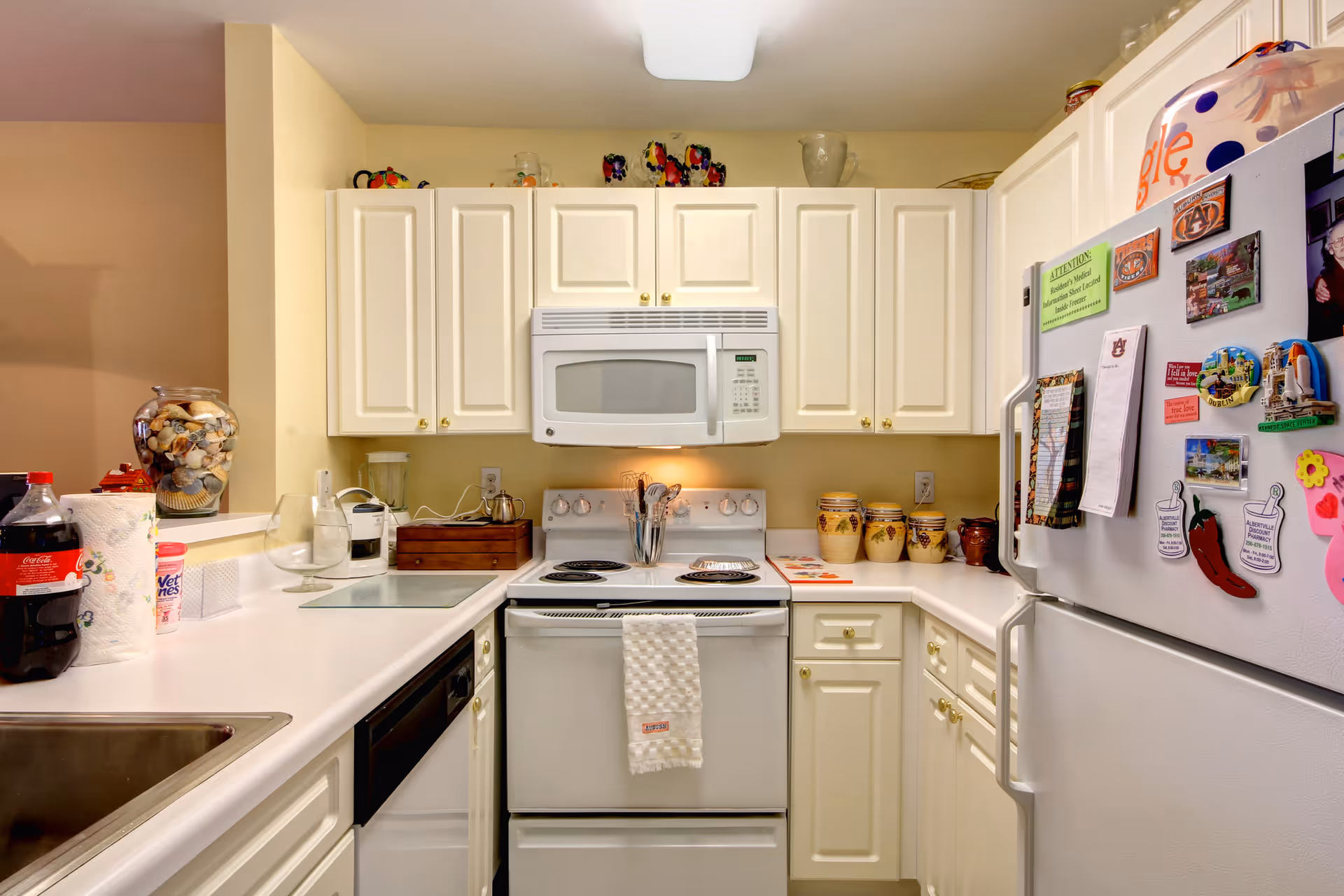 A compact kitchen with white cabinets and appliances including a microwave, stove, dishwasher, and refrigerator. The countertops hold various items such as a Coca-Cola bottle, paper towels, a blender, a glass bowl, and decorative jars. The refrigerator door is covered with magnets and notes.
