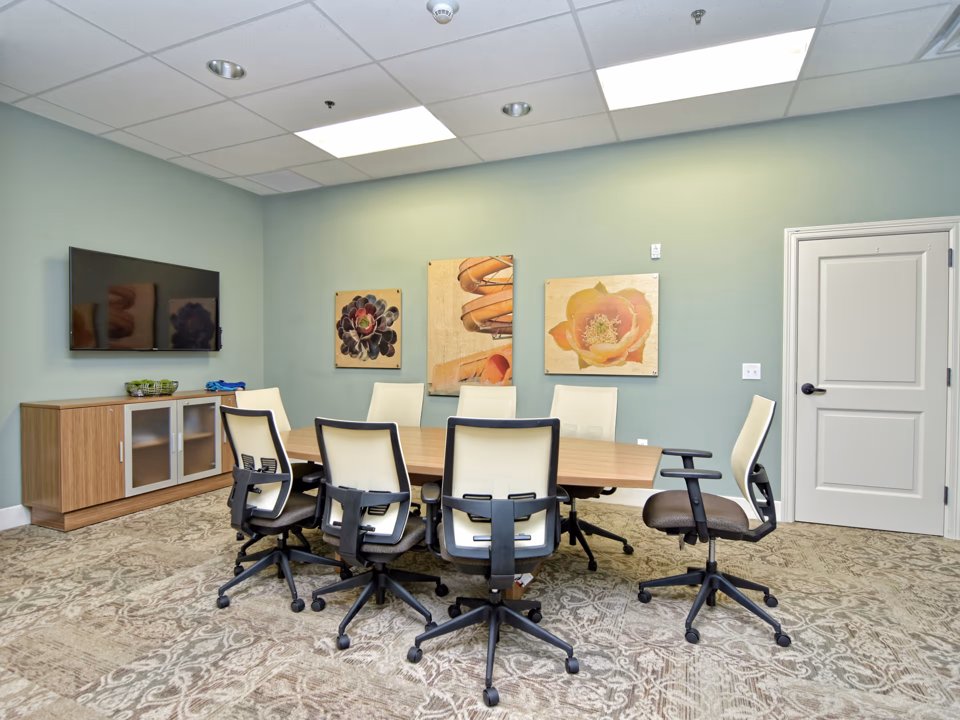 A conference room with a rectangular wooden table surrounded by eight office chairs with white backs and black frames. On the wall behind the table are three framed artworks featuring floral and abstract designs. A flat-screen TV is mounted on the left wall above a wooden cabinet with frosted glass doors. The room has a patterned carpet and a white door on the right side.