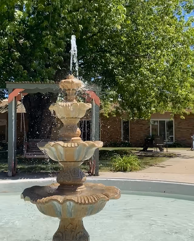 Ornate tiered fountain spraying water in a sunny courtyard with trees and a brick building in the background.