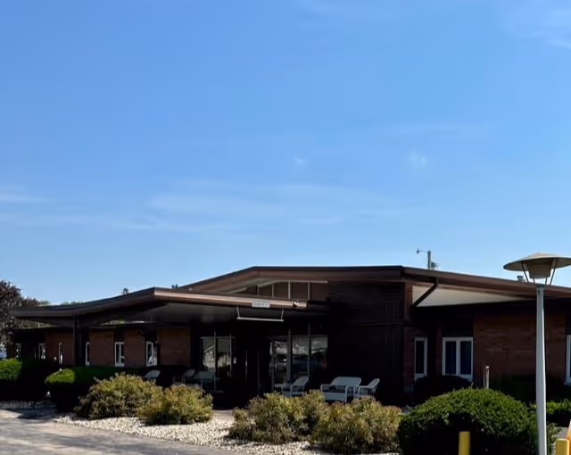 Single-story brick building front with a covered entrance, outdoor seating, and landscaped shrubs under a clear blue sky.