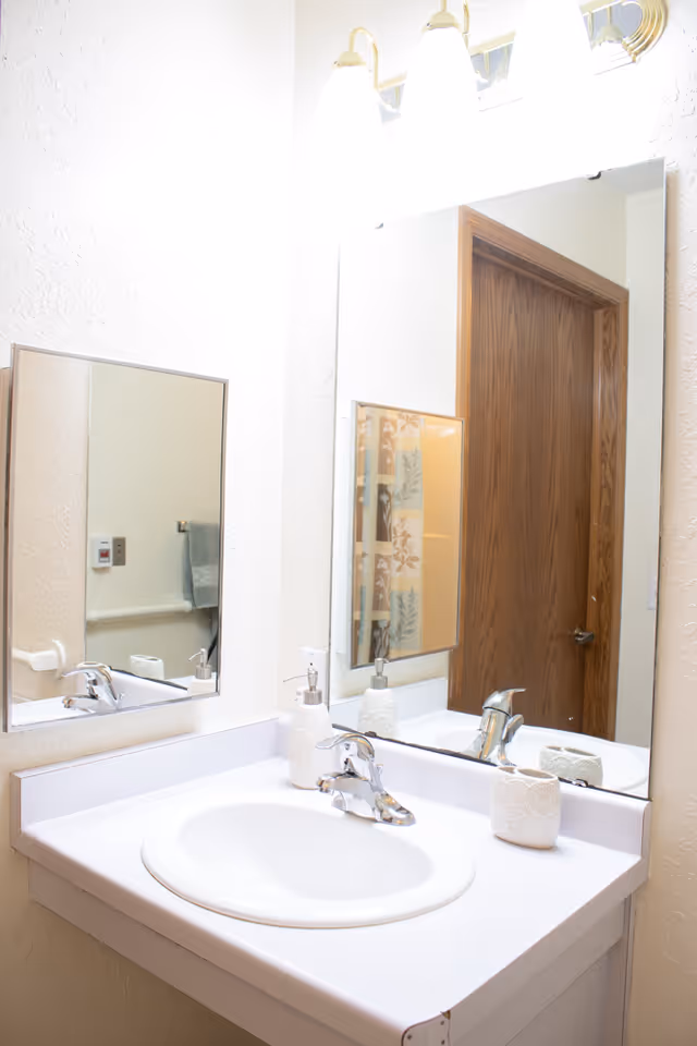 Bathroom vanity with a white sink and countertop, chrome faucet, soap dispenser and a large mirror reflecting a wooden door.