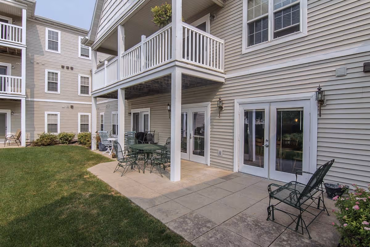 Outdoor patio area at The Woodlands Senior Apartments featuring a concrete patio with metal chairs and tables, adjacent to a beige multi-story building with white trim and balconies. There is a grassy lawn beside the patio and some small bushes and flowers near the building.