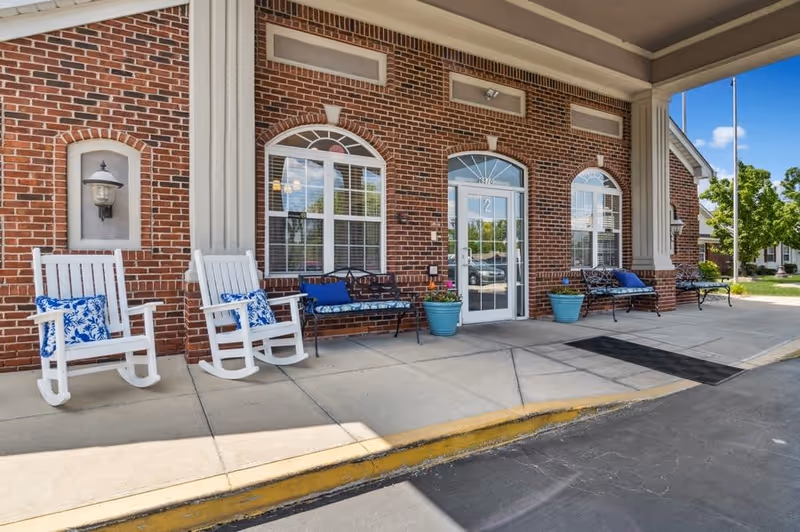 Entrance area of a brick building with white-framed windows and a glass door. There are two white rocking chairs with blue and white patterned cushions, two black metal benches with blue cushions, and potted plants placed along the wall. The ground is paved with concrete and there is a yellow curb along the edge.