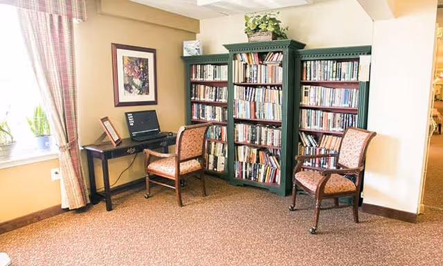 A cozy reading and computer area in a senior living facility with two wooden chairs upholstered in patterned fabric, a small dark wooden desk with a laptop, and two green bookshelves filled with books. A framed floral picture hangs on the wall above the desk, and a window with plaid curtains lets in natural light.