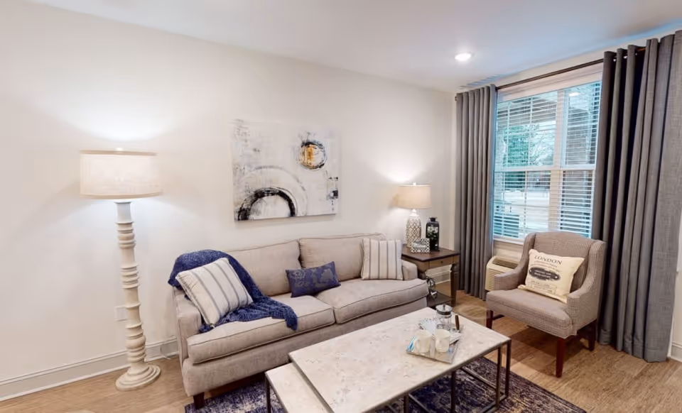 A cozy living room with a beige sofa adorned with striped and patterned pillows, a blue throw blanket, a cushioned armchair with a decorative pillow, a marble coffee table with cups and a French press, two table lamps on either side of the sofa, and a large window with gray curtains letting in natural light.