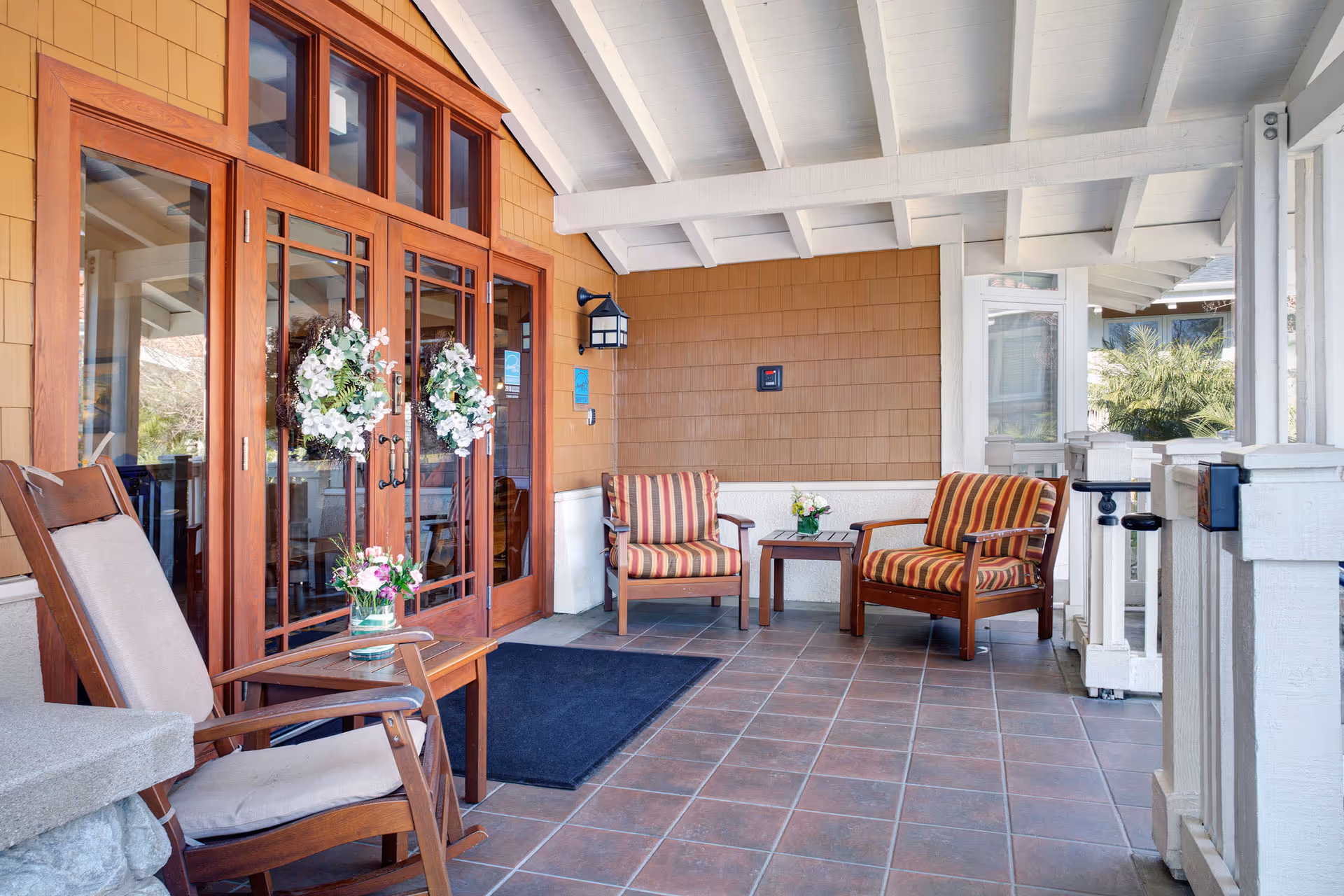 Covered porch area with wooden rocking chairs and cushioned armchairs arranged around small tables. The porch features a tiled floor, wooden framed glass doors decorated with white floral wreaths, and a white beamed ceiling. There are flowers in vases on the tables and a wall-mounted lantern light fixture.