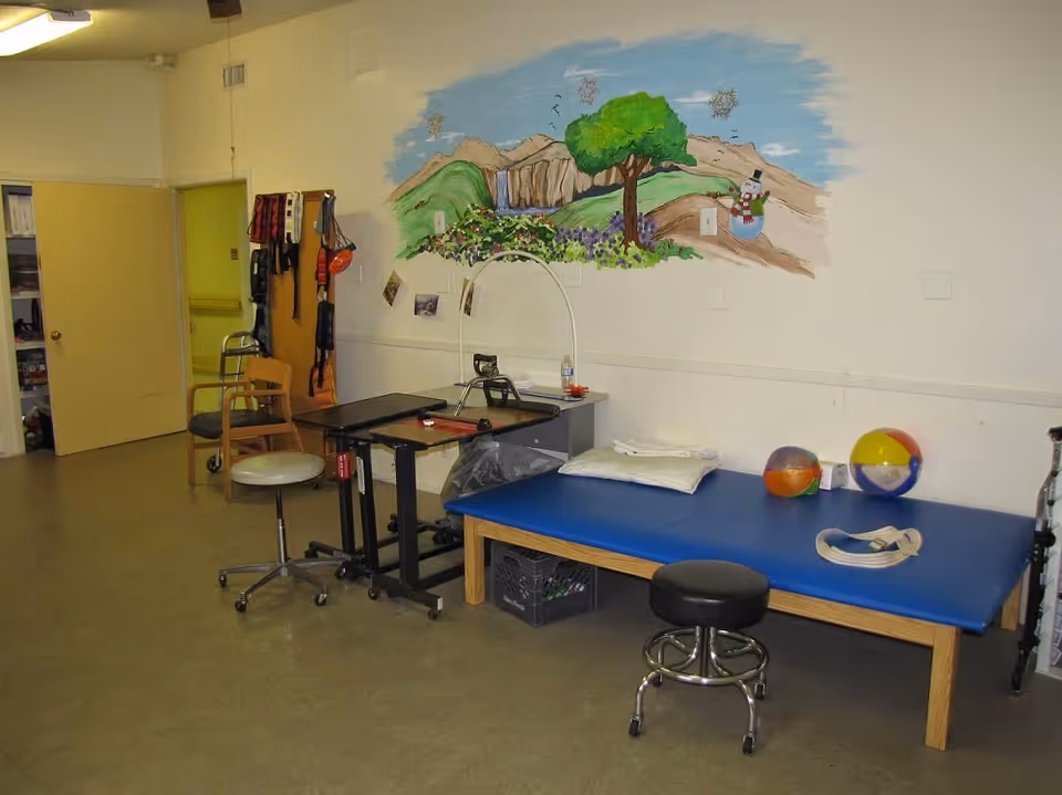 A therapy or rehabilitation room with a blue padded treatment table, two stools on wheels, a small black adjustable table, and a wooden chair. The wall features a colorful mural of a tree, mountains, and a snowman. Various therapy equipment and accessories are visible, including a walker and exercise bands hanging on a wooden rack.