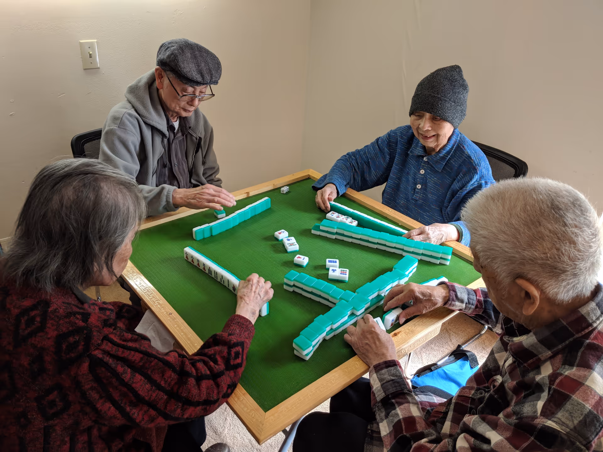 Four elderly individuals sitting around a green mahjong table playing a game. They are focused on the game pieces, with two men and two women participating. The room has plain beige walls and simple chairs.
