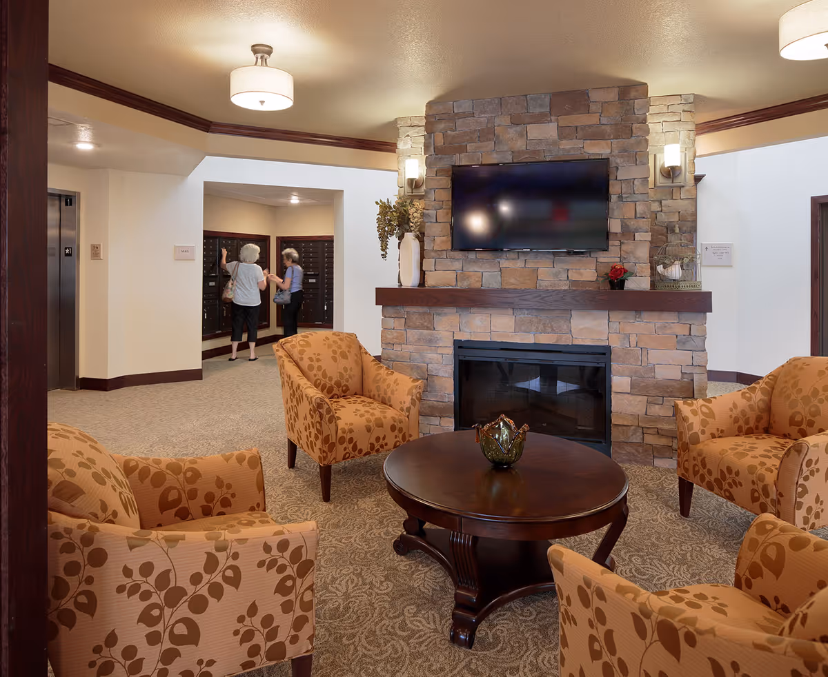 A cozy common area in a senior living facility with four patterned armchairs arranged around a round wooden coffee table. A stone fireplace with a mounted flat-screen TV is the focal point of the room. In the background, two elderly women are seen near a bank of mailboxes. The room has warm lighting and neutral-colored walls and carpet.
