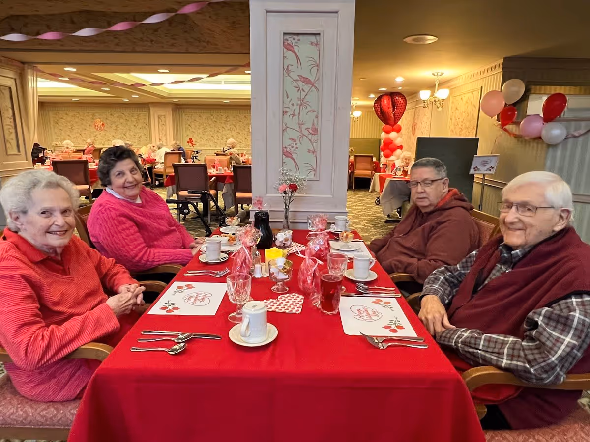 Four elderly people sitting around a table covered with a red tablecloth in a dining room decorated with Valentine's Day themed items including heart-shaped balloons, pink and red streamers, and placemats with hearts. The room has other tables with elderly residents in the background.