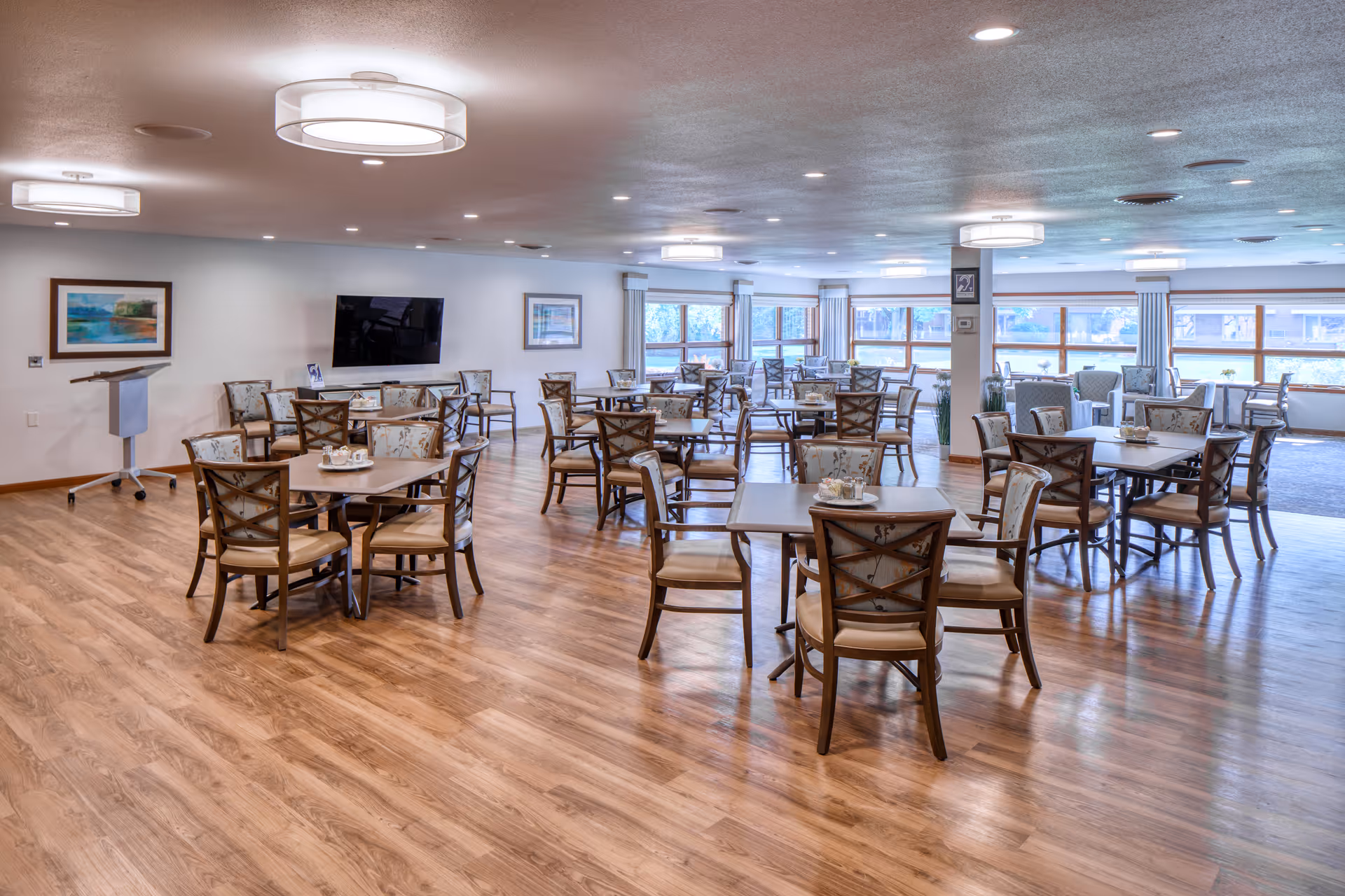 A spacious dining room with multiple wooden tables and chairs arranged neatly on a wood floor. The room has large windows letting in natural light, framed artwork on the walls, ceiling lights, and a mounted flat-screen TV.
