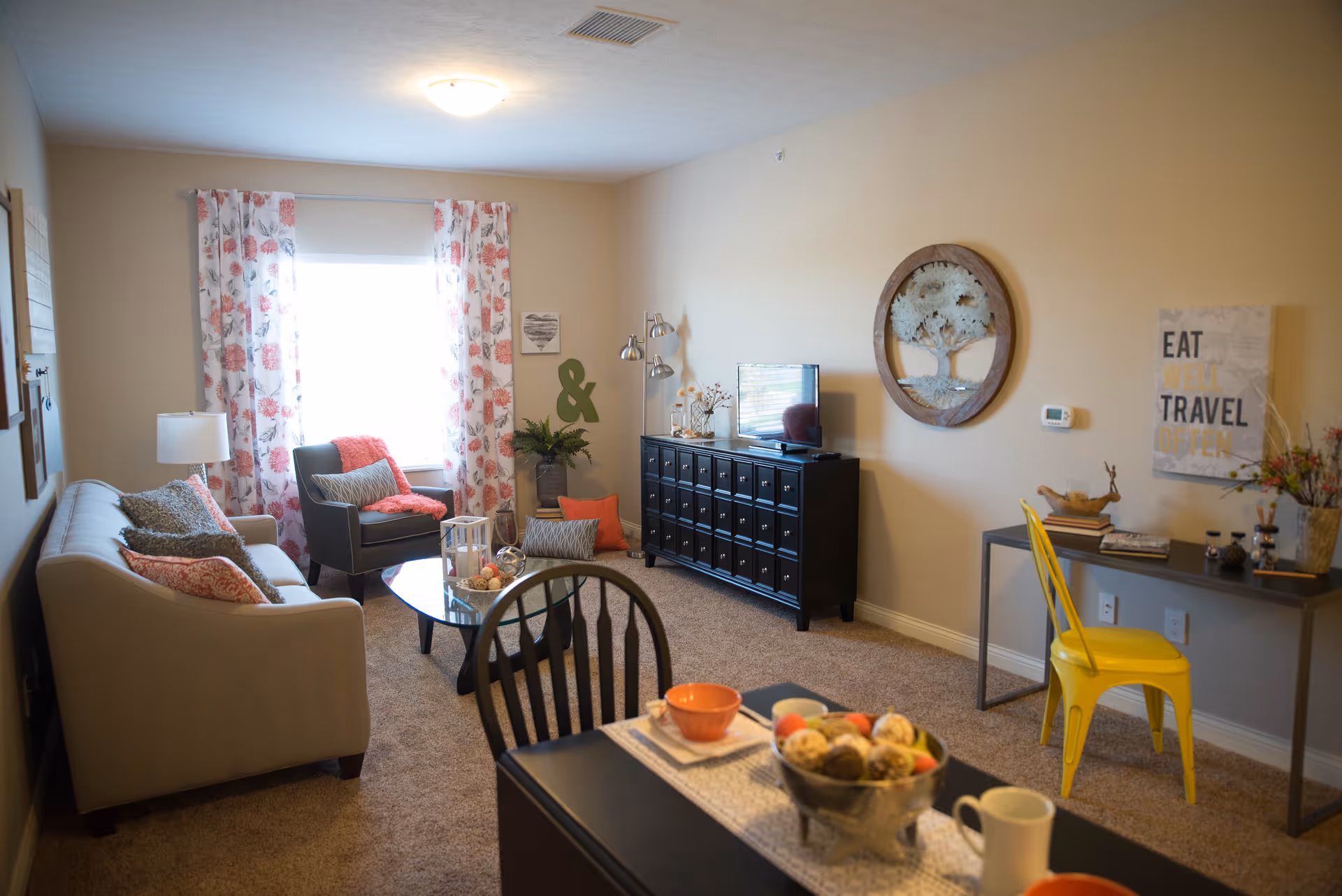 A cozy living room with a beige sofa adorned with patterned pillows, a gray armchair with a coral throw, and a round glass coffee table. The room has floral curtains on the window, a black cabinet with a TV on top, and a wall decoration of a tree. There is a small desk with a yellow chair and decorative items, including a sign that says 'EAT TRAVEL'. The room is carpeted and softly lit by a ceiling light.