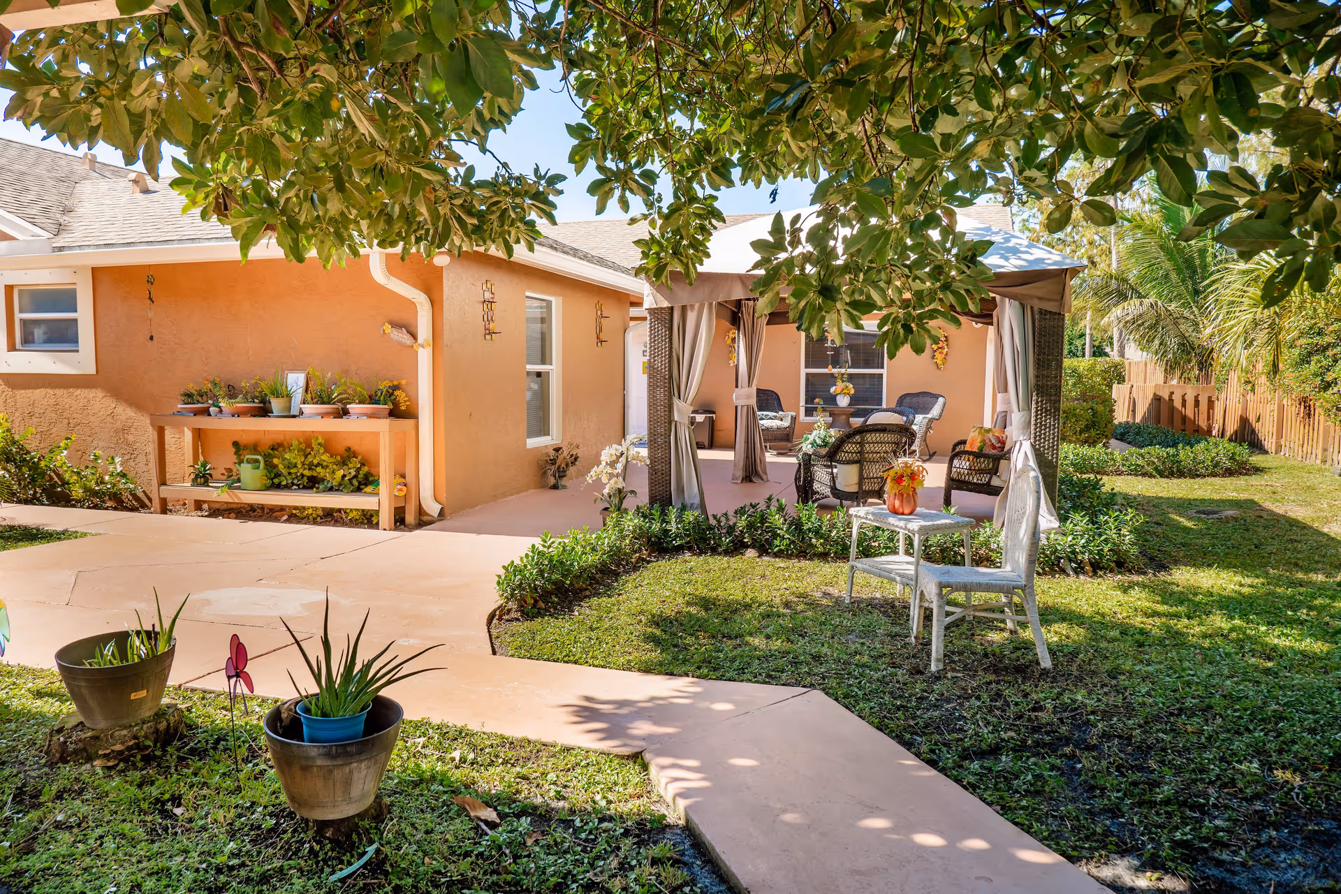 Outdoor patio area at The Club at Wellington Assisted Living & Memory Care featuring a paved walkway, green lawn with potted plants, a small white table with a chair, and a shaded seating area with wicker chairs and curtains under a canopy. The building exterior is light brown with windows and decorative plants.