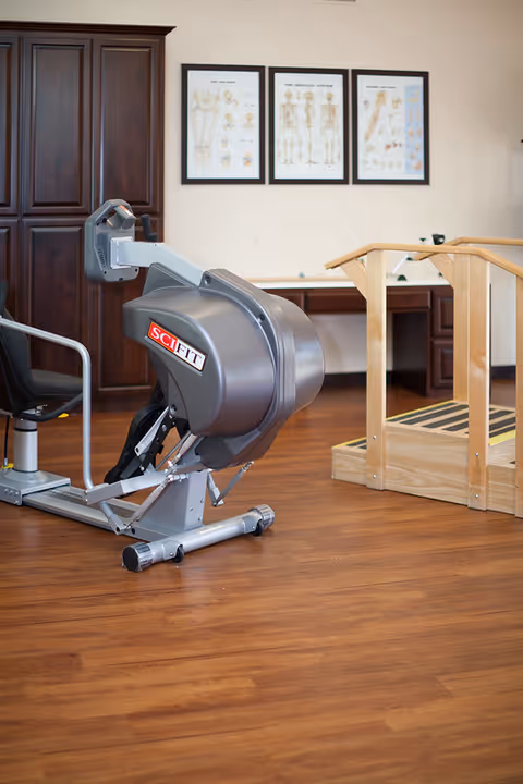 A rehabilitation or physical therapy room with a SciFit exercise machine, wooden parallel bars for walking practice, a dark wooden cabinet, and anatomical charts on the wall.