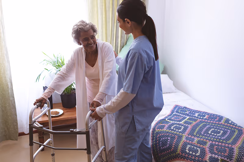 A caregiver assists an elderly woman using a walker beside a bed in a sunny bedroom.