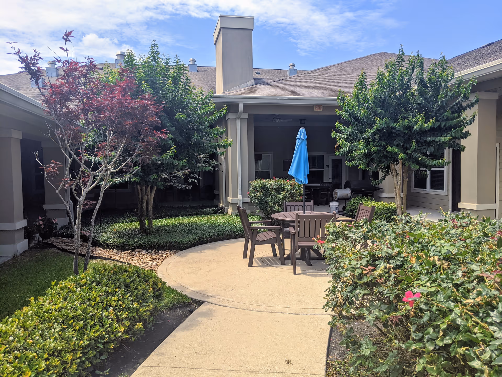 Outdoor courtyard area at Orchard Park at Victory Lakes Assisted & Senior Living featuring a circular patio with a round table, several chairs, and a blue umbrella. The courtyard is surrounded by green bushes, trees, and a building with beige walls and a shingled roof under a partly cloudy sky.
