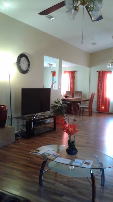Open-plan living room and dining area with a TV on a stand, glass coffee table with a floral arrangement, hardwood floors, and red curtains.