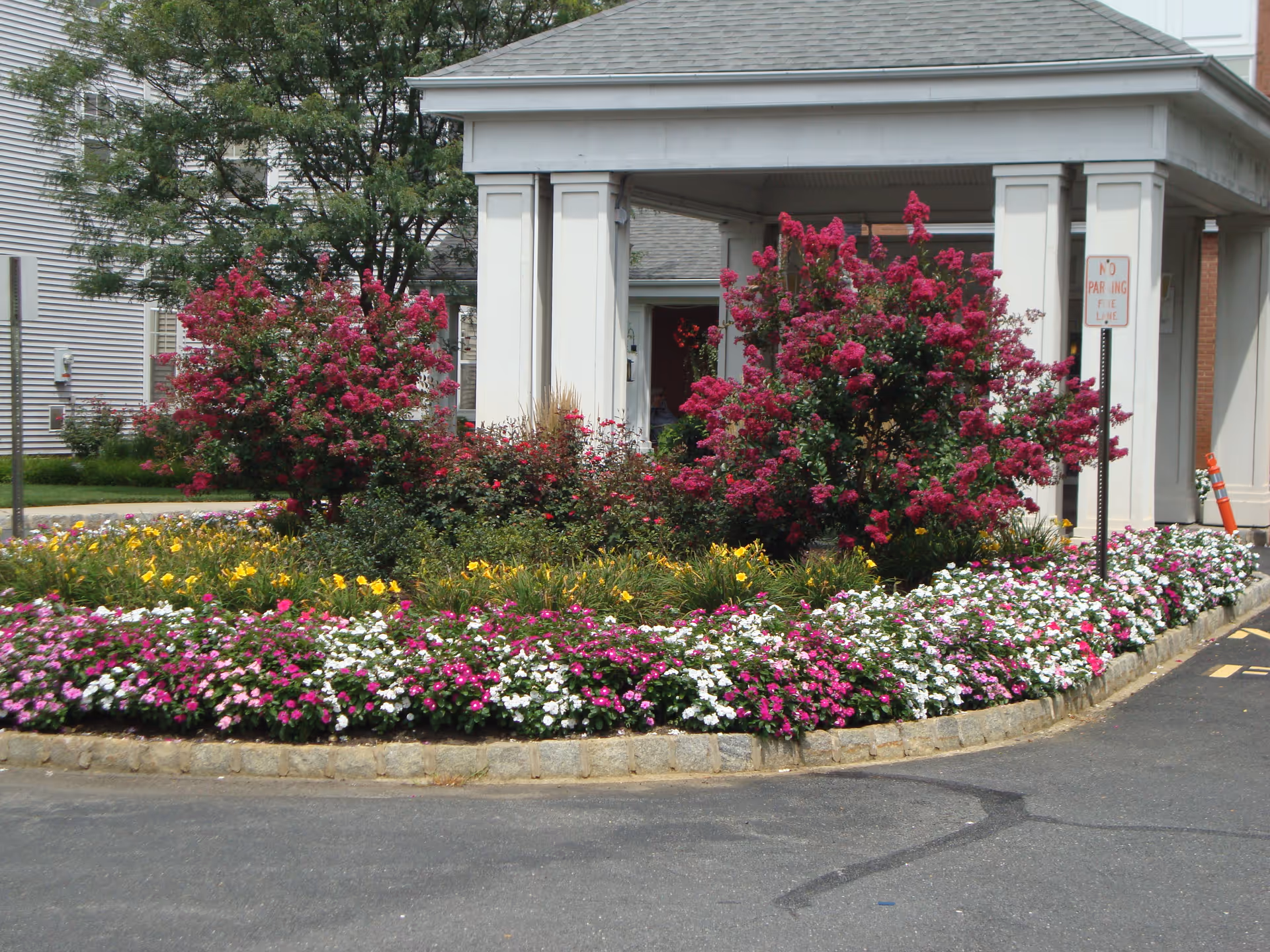 A landscaped garden area with vibrant pink, white, and yellow flowers in front of a white building entrance with columns. There is a 'No Parking Fire Lane' sign visible near the entrance.