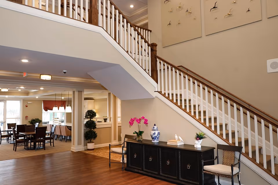 Interior view of a senior living facility showing a staircase with wooden handrails and white balusters. Below the staircase is a black console table decorated with a vase of pink flowers, a blue and white ceramic jar, books, and a small plant. Two chairs with cushions are placed on either side of the table. In the background, there is a dining area with tables and chairs, pendant lights, and large windows allowing natural light to enter.
