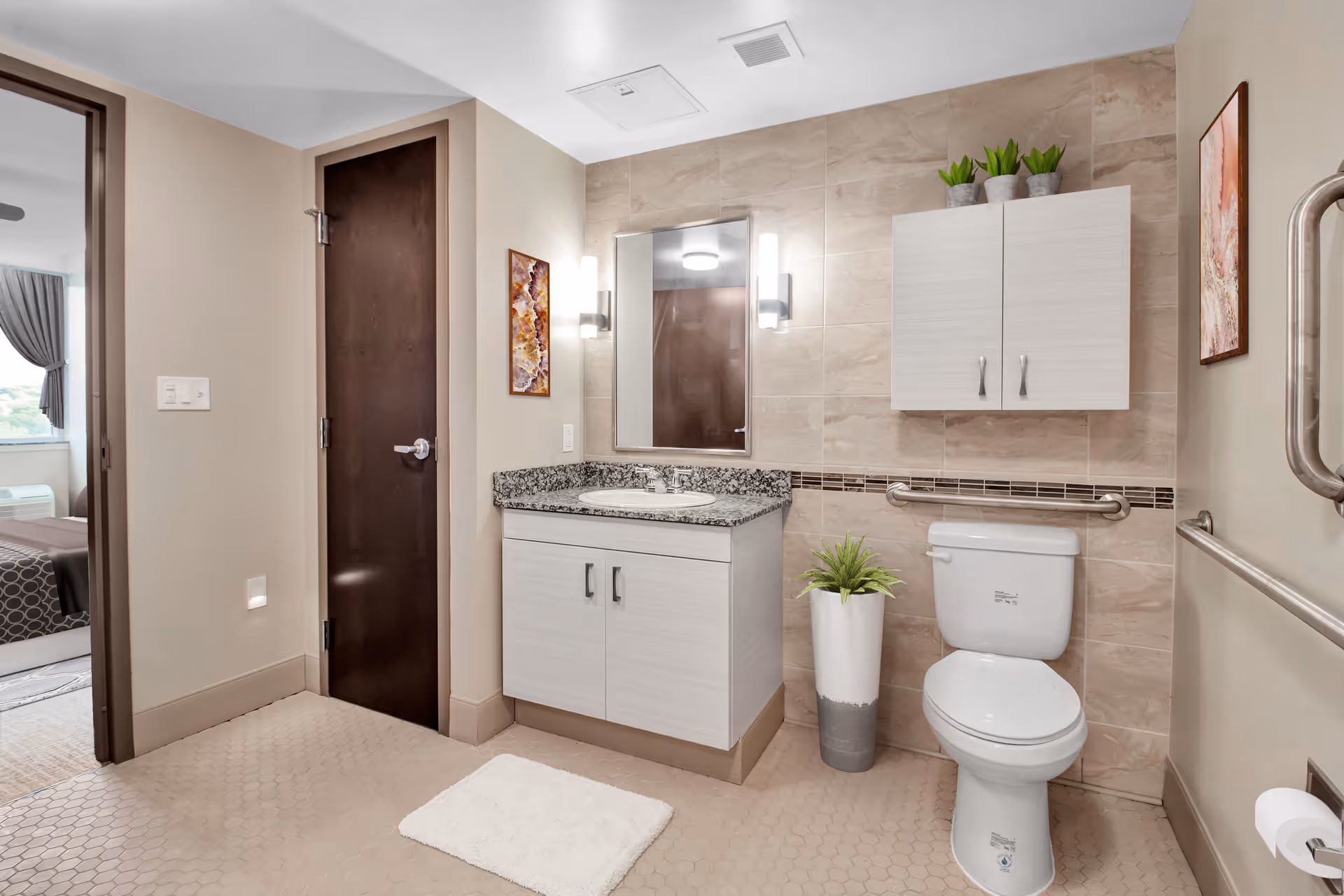 A clean and modern bathroom with beige tiled walls and hexagonal floor tiles. The room features a white toilet with a grab bar beside it, a white vanity with a granite countertop and a sink, a mirror with two vertical lights on either side, a wall-mounted cabinet with potted plants on top, and a small potted plant on the floor. A dark wooden door leads to an adjacent bedroom visible through the doorway.