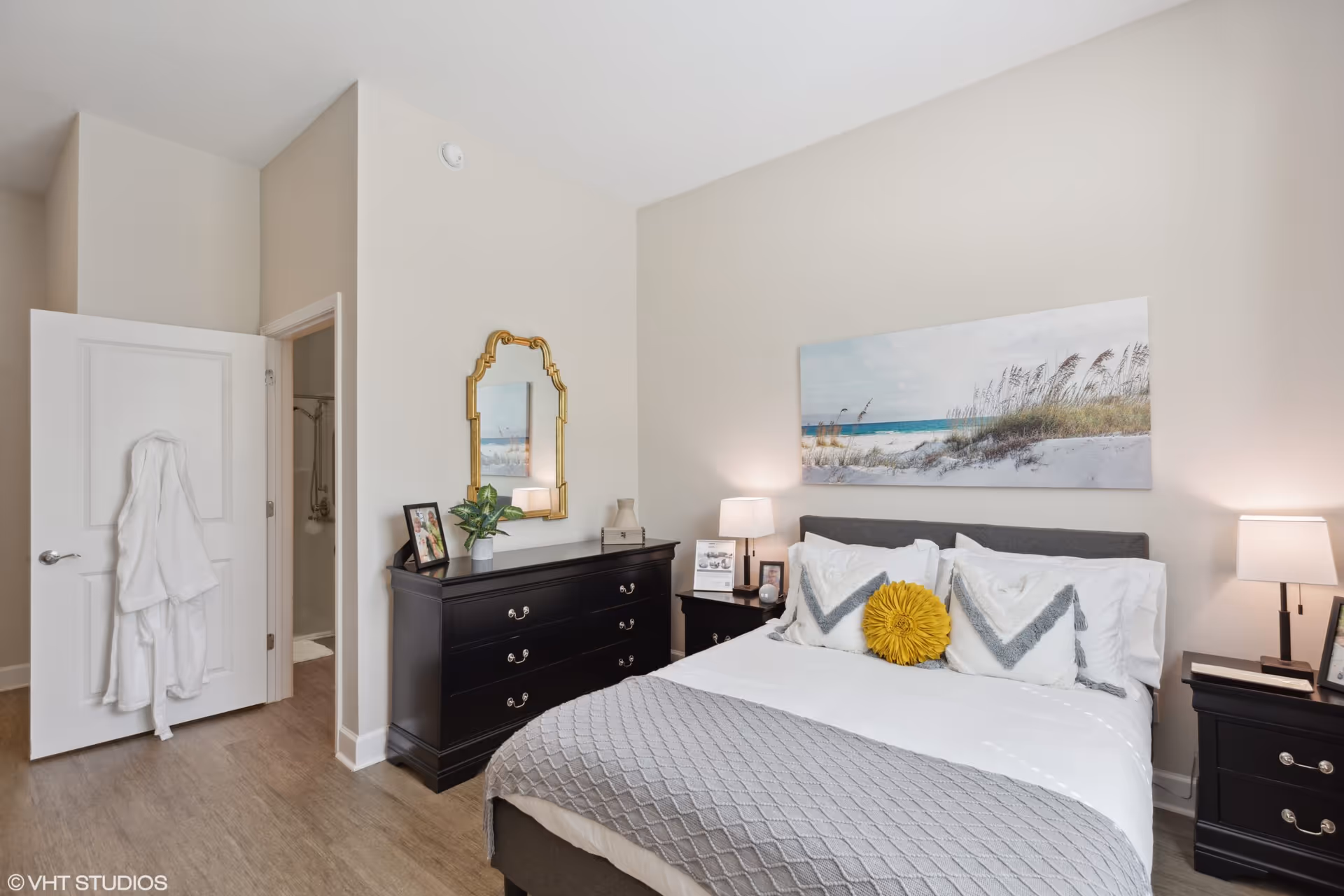 Bright, modern bedroom with a neatly made bed, two nightstands with lamps, a dresser topped with a mirror and decor, and an open door leading to a bathroom.