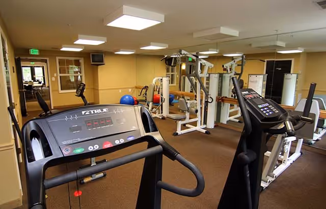 Interior view of a fitness room with exercise equipment including treadmills, weight machines, and exercise balls. The room has beige walls, brown carpet, and ceiling lights. Mirrors cover one wall, reflecting the equipment and space.