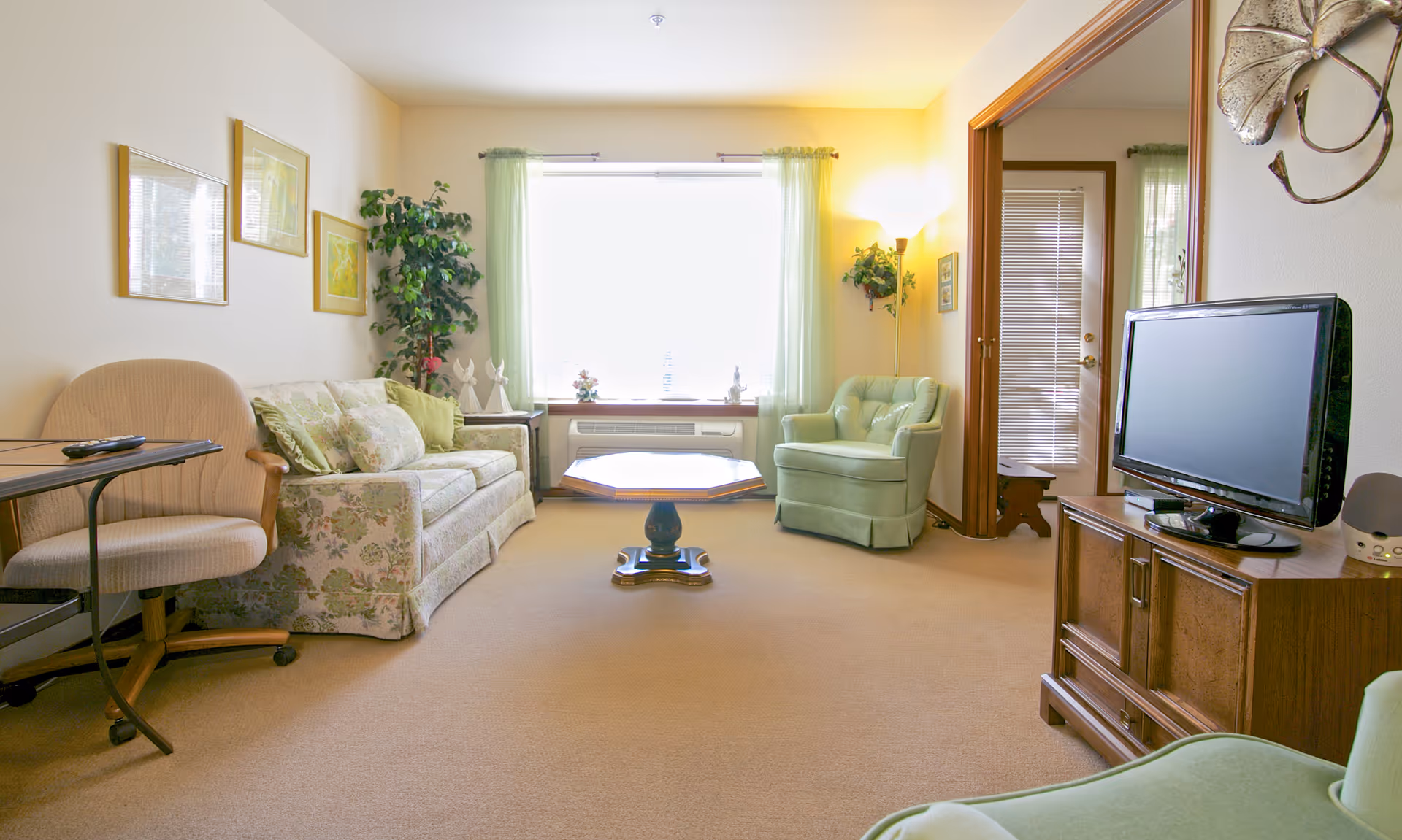 A cozy living room with a floral patterned sofa, a green armchair, a wooden coffee table, a TV on a wooden cabinet, and a window with light green curtains letting in bright natural light. The room has beige carpet and light-colored walls decorated with framed pictures and plants.