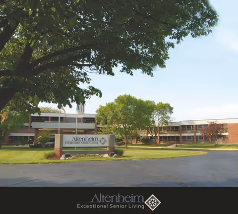 Exterior view of Altenheim Family-first Senior Living facility showing a two-story brick building with large windows, surrounded by green trees and a well-maintained lawn. A sign in front reads 'Altenheim Exceptional Senior Living'.