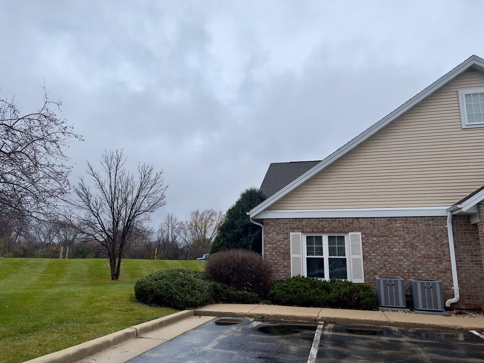 Exterior view of a building with beige siding and brick walls, two windows with white shutters, and two air conditioning units next to the building. In front of the building is a parking area with wet pavement and a grassy area with bushes and leafless trees under a cloudy sky.
