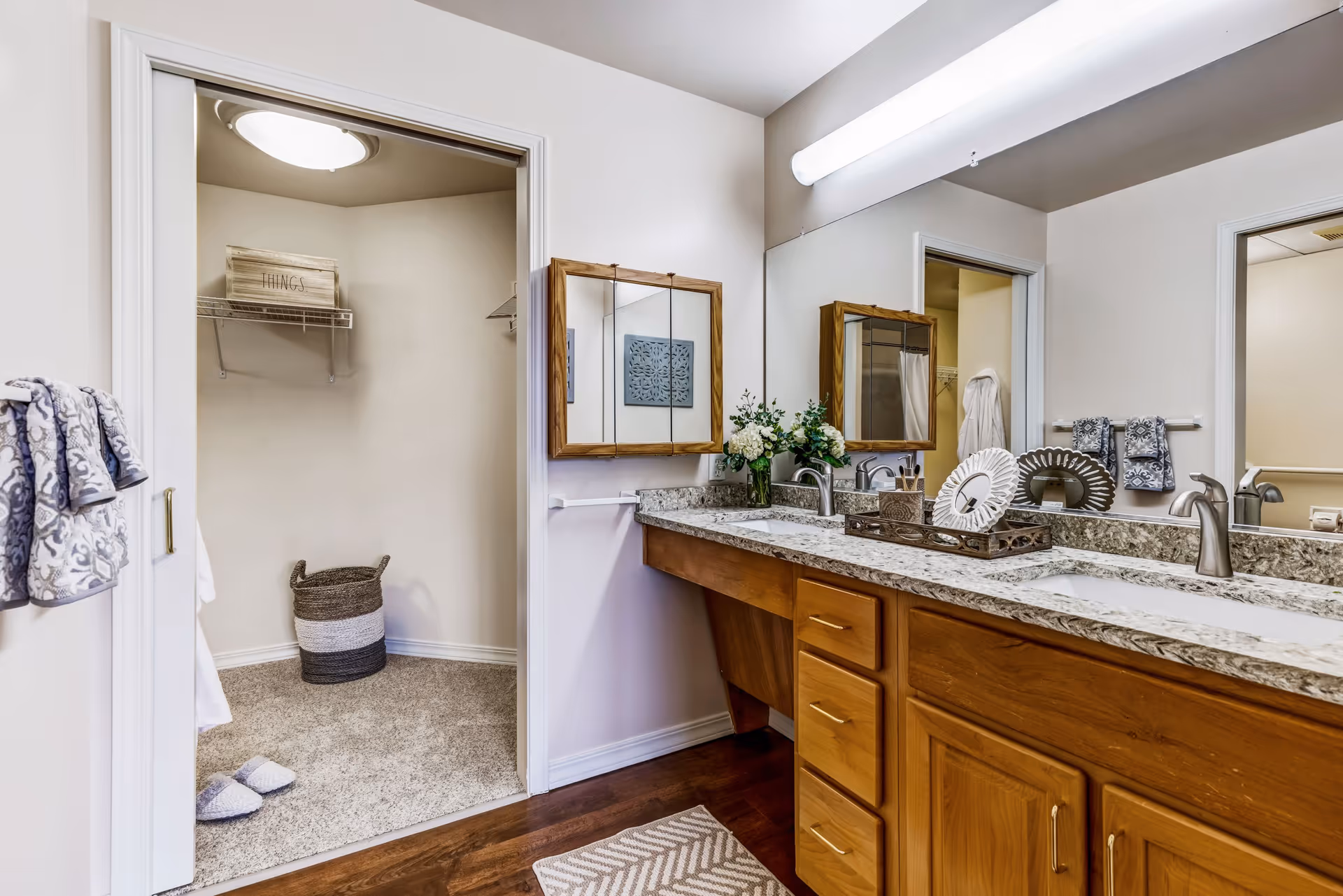 A bathroom with a double sink vanity featuring granite countertops and wooden cabinets. Above the sinks are two wooden framed mirrors and a large horizontal light fixture. On the countertop, there are decorative items including a vase with flowers and a tray with ornamental pieces. To the left, there is an open doorway leading to a carpeted walk-in closet with a shelf labeled 'THINGS' and a woven basket on the floor. Towels hang on the wall and a patterned rug is on the wooden floor in front of the vanity.