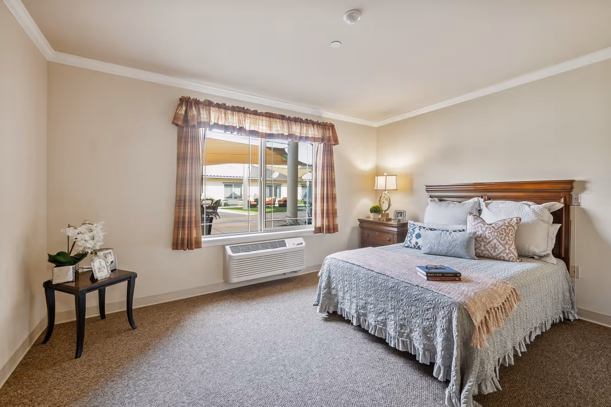 A cozy bedroom in an assisted living facility featuring a neatly made bed with multiple pillows and a light blue bedspread. There is a wooden nightstand with a lamp and decorative items beside the bed. A small table with a potted plant and framed photos is placed near a window with plaid curtains. Outside the window, a patio area with chairs and tables is visible.