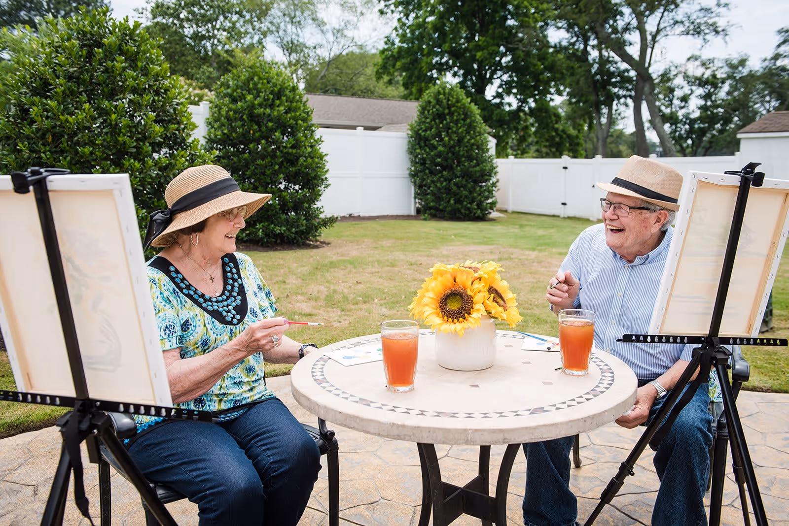 Two elderly individuals sitting outdoors at a round table with a vase of sunflowers and two glasses of iced tea, each painting on canvases set on easels, smiling and enjoying the activity in a fenced garden area.