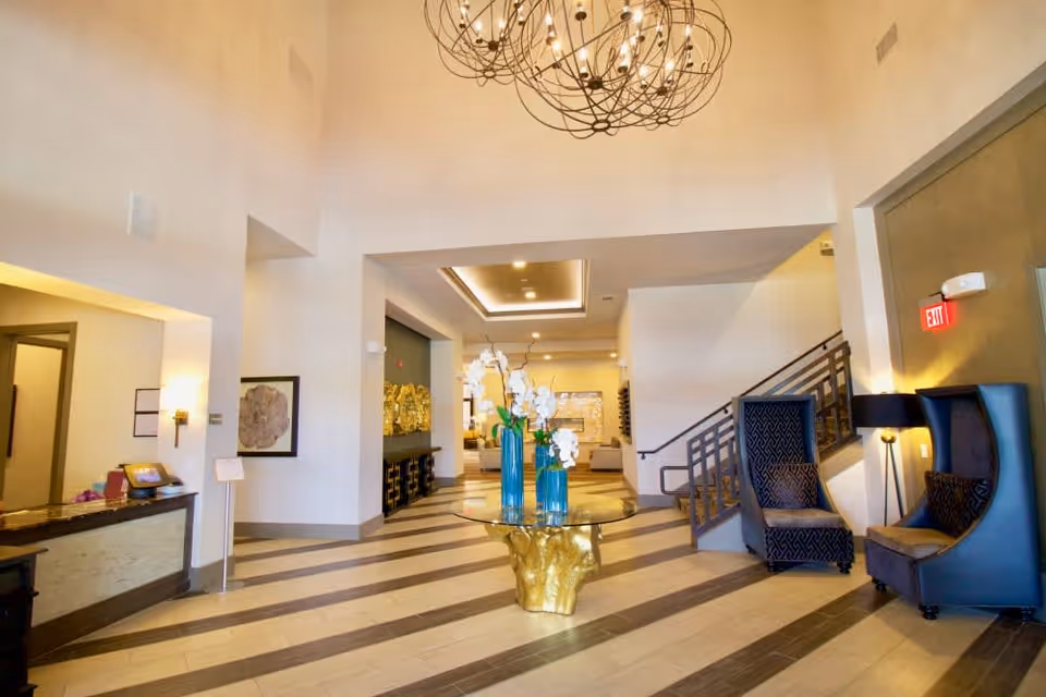 Spacious and well-lit lobby area with a high ceiling, featuring a round glass table with a gold base and blue vases with white flowers in the center. To the right, there are two high-backed blue armchairs with patterned cushions next to a floor lamp. On the left side, there is a reception desk with a computer and some items on it. The floor has a striped pattern, and a modern spherical chandelier hangs from the ceiling. A staircase is visible on the right side, and the overall decor is modern and elegant.
