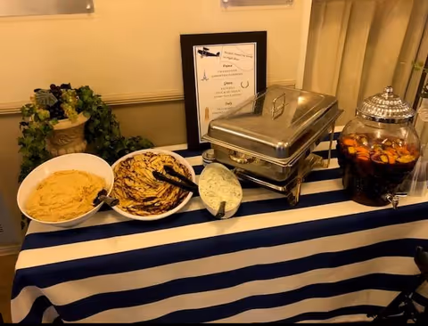 A buffet table with bowls of dips, a chafing dish, and a glass beverage dispenser on a blue-and-white striped tablecloth.