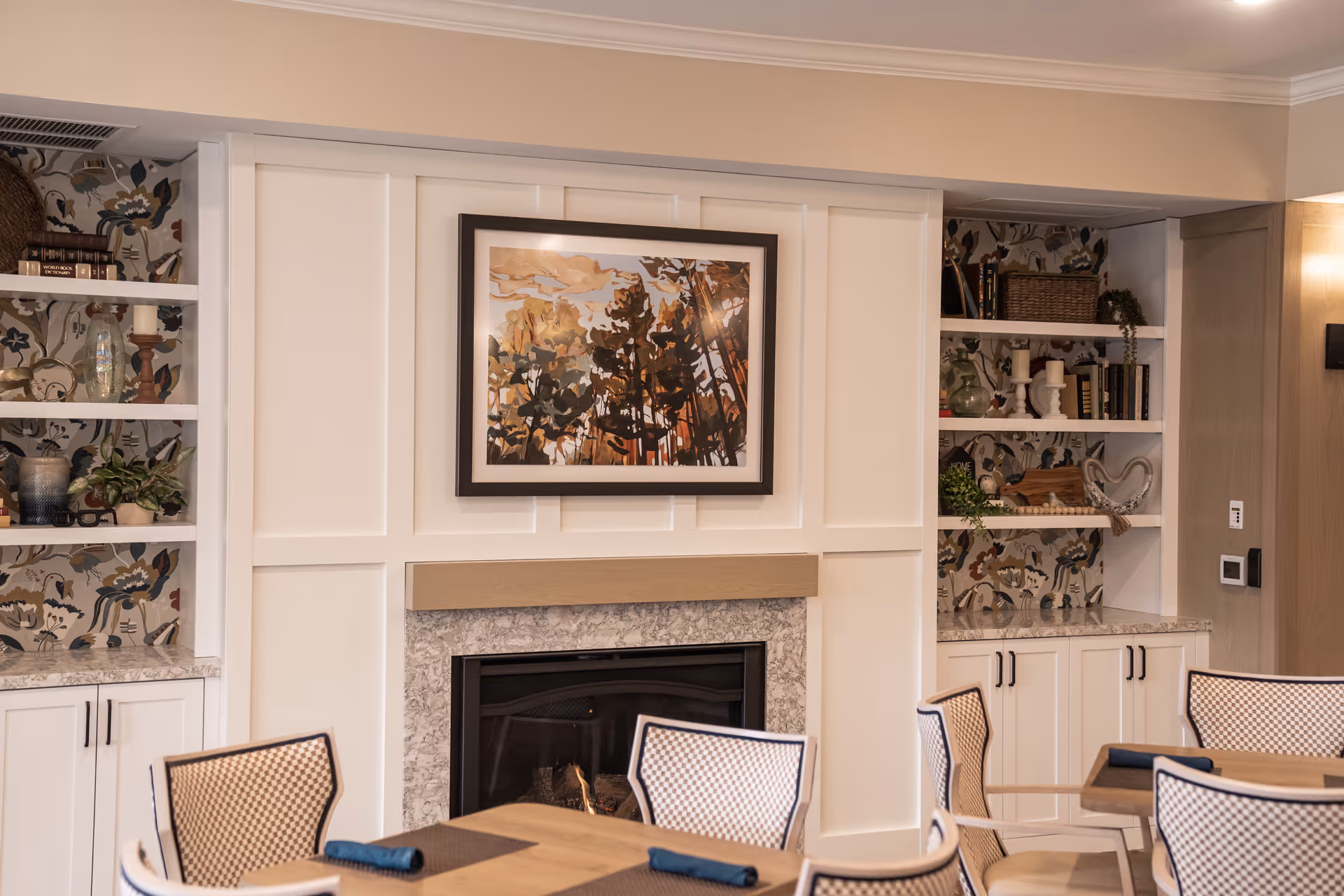 Dining area with tables and chairs facing a fireplace flanked by built-in shelves and a framed artwork above.