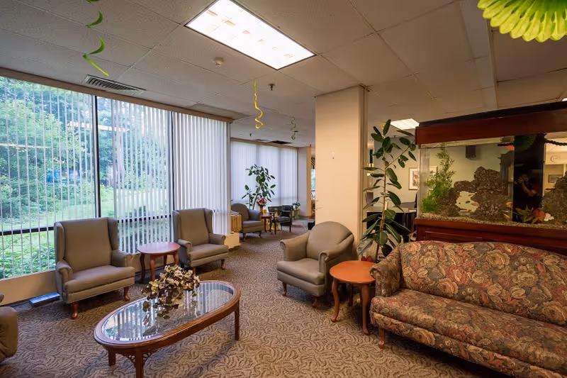 A bright senior living facility sitting area with upholstered chairs, a patterned sofa, coffee tables, large windows with vertical blinds, plants and an aquarium.