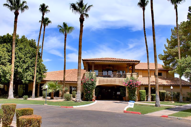 Exterior view of a two-story building with a tiled roof and a covered entrance. The building is surrounded by tall palm trees, green bushes, and a well-maintained lawn under a partly cloudy blue sky.