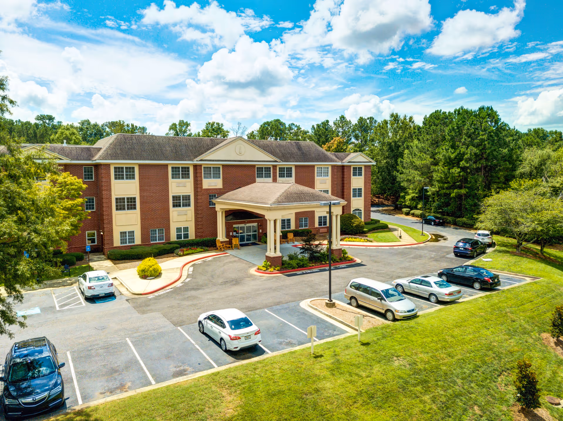 Exterior view of a three-story brick building with a covered entrance and several parked cars in front. The building is surrounded by green trees and grass under a partly cloudy blue sky.
