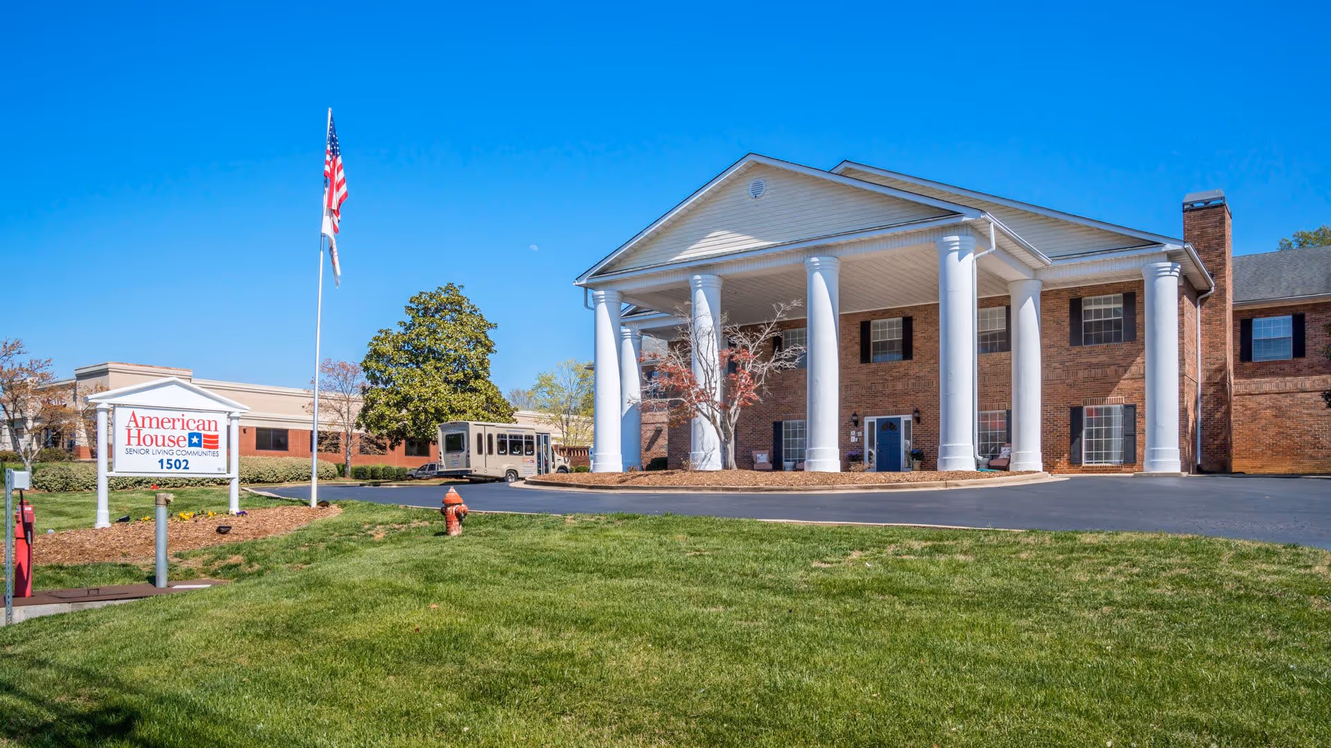 Exterior view of American House Hamilton Place senior living community building with large white columns at the entrance, a green lawn in front, an American flag on a flagpole, and a shuttle bus parked near the entrance under a clear blue sky.