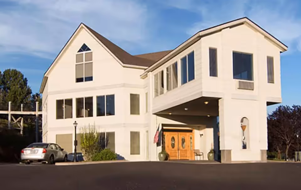 White multi-story building with large windows and a covered front entrance, a parked car nearby, and a clear sky.